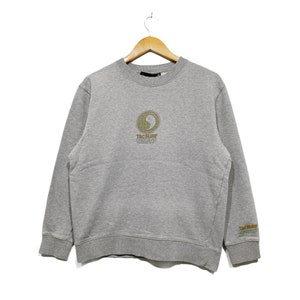 May include: A heather gray sweatshirt with a crew neck and long sleeves. The front features a circular design with the text "T&C Surf Designs". The same text is repeated on the right sleeve. The sweatshirt is hanging on a wooden hanger.