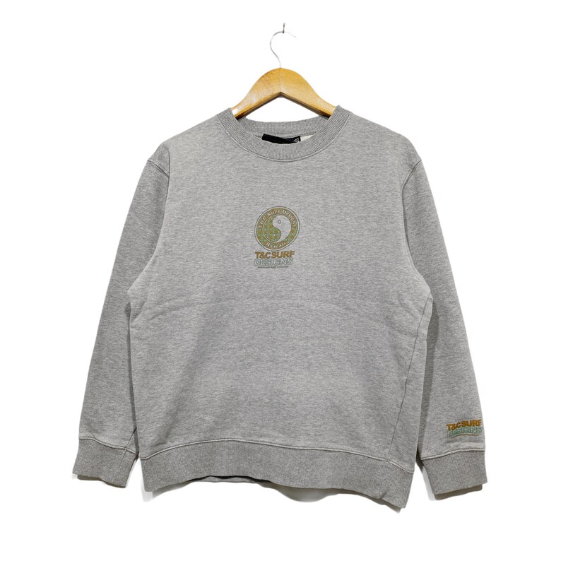 May include: A heather gray sweatshirt with a crew neck and long sleeves. The front features a circular design with the text "T&C Surf Designs". The same text is repeated on the right sleeve. The sweatshirt is hanging on a wooden hanger.