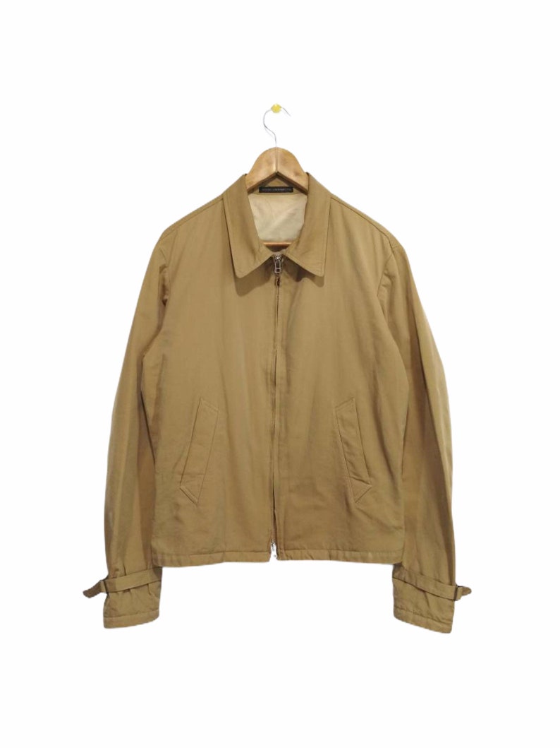 May include: A tan zip-up jacket with long sleeves and a collared neckline. The jacket has a strap detail on the cuffs.