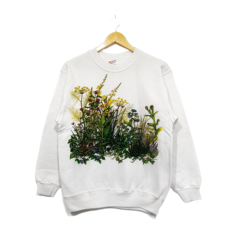 Op de afbeelding: Witte trui met een bloemenprint van verschillende bloemen en groen. De print is in tinten groen, geel, roze en blauw.