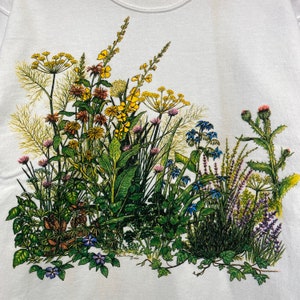 Op de afbeelding: Een wit T-shirt met een kleurrijke bloemenprint met gele, blauwe, paarse en groene bloemen. De bloemen zijn in een cluster gerangschikt en lijken in een veld te groeien.