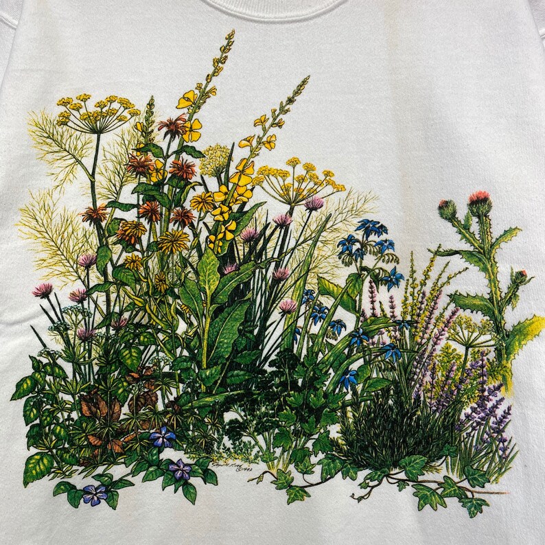 Op de afbeelding: Een wit T-shirt met een kleurrijke bloemenprint met gele, blauwe, paarse en groene bloemen. De bloemen zijn in een cluster gerangschikt en lijken in een veld te groeien.