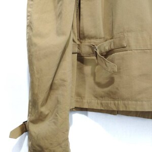 May include: A tan utility jacket with a long sleeve and a belt. The jacket has a flap pocket on the back and a buckle closure on the sleeve.