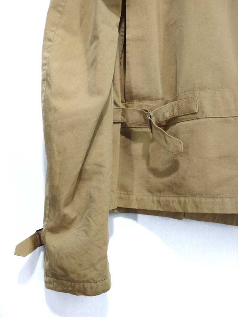 May include: A tan utility jacket with a long sleeve and a belt. The jacket has a flap pocket on the back and a buckle closure on the sleeve.