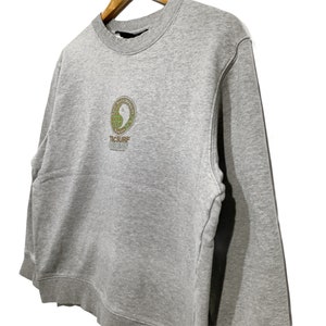 May include: A gray crew neck sweatshirt with long sleeves. The front features a circular design with a yin-yang symbol and the text "T&C Surf Designs Hawaii". The sweatshirt is hanging on a wooden hanger.