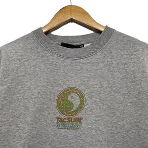 May include: A gray sweatshirt with a crew neck, hanging on a wooden hanger. The front features a circular design with a yin-yang symbol and the text "T&C Surf Designs Hawaii". The design is in shades of green and brown.