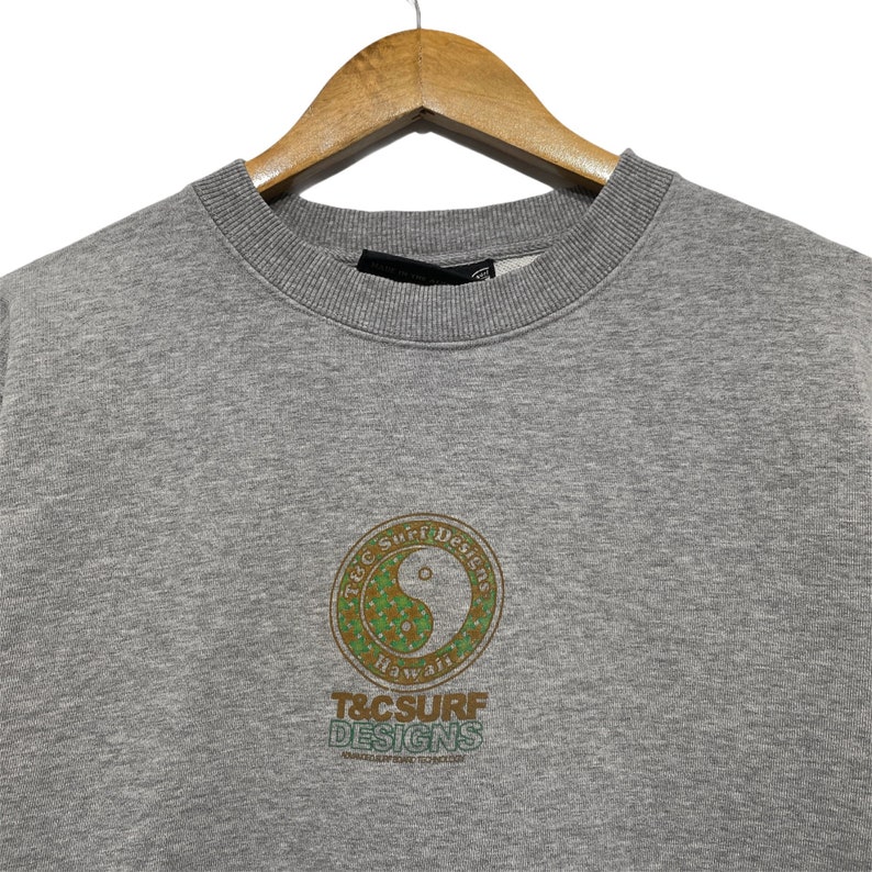 May include: A gray sweatshirt with a crew neck, hanging on a wooden hanger. The front features a circular design with a yin-yang symbol and the text "T&C Surf Designs Hawaii". The design is in shades of green and brown.