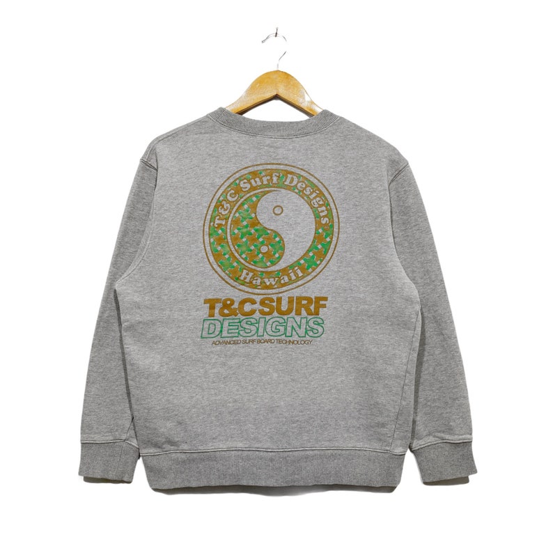 May include: A heather gray sweatshirt with a round neck and long sleeves. The back features a circular design with a yin-yang symbol and the text "T&C Surf Designs Hawaii". Below the design is the brand name in green and white.