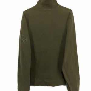 May include: A green long-sleeved shirt with a high neck and a compass logo on the left sleeve.