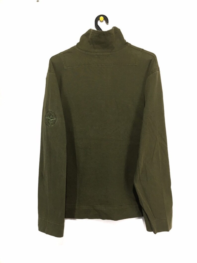 May include: A green long-sleeved shirt with a high neck and a compass logo on the left sleeve.