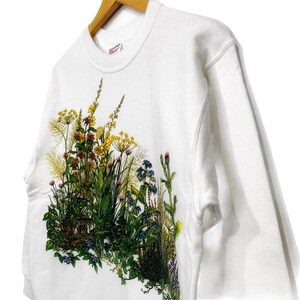 Op de afbeelding: Witte trui met een bloemenprint met verschillende bloemen en bladeren in tinten groen, geel, blauw en roze.