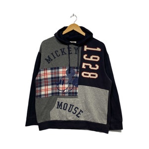 Puede incluir: Una sudadera con capucha de color azul, gris y rojo con un gr&aacute;fico de Mickey Mouse y el texto "Mickey Mouse 1928" en la parte delantera.