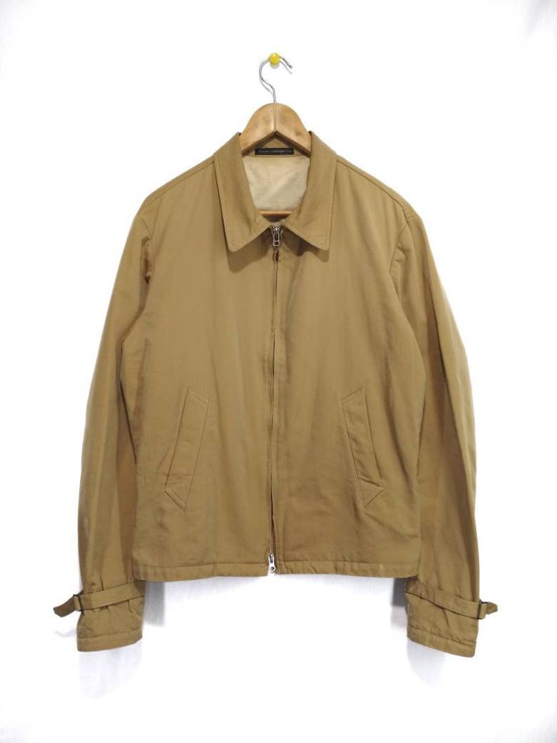 May include: A tan zip-up jacket with long sleeves and a collared neckline. The jacket has a zipper closure and two pockets on the front. The sleeves have adjustable straps at the cuffs.