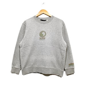 May include: A heather gray sweatshirt with a crew neck and long sleeves. The front features a circular design with a yin-yang symbol and the text "T&C Surf Designs". The right sleeve has the text "T&C Surf Designs" in green.