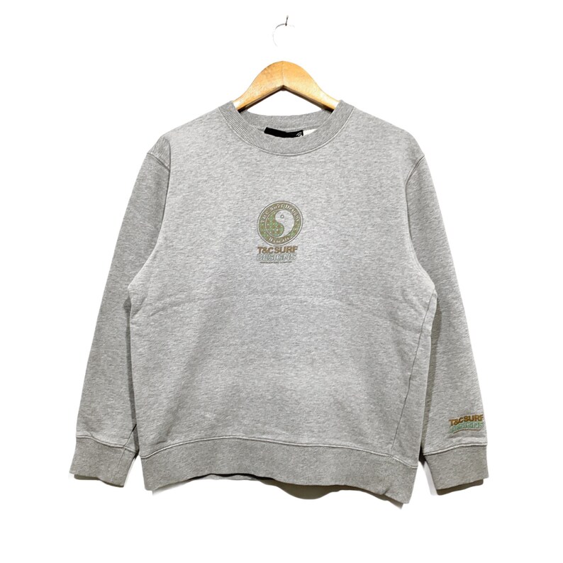 May include: A heather gray sweatshirt with a crew neck and long sleeves. The front features a circular design with a yin-yang symbol and the text "T&C Surf Designs". The right sleeve has the text "T&C Surf Designs" in green.