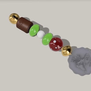 Può includere: Una collezione di perline e ciondoli decorativi in varie forme e colori. Include sfere dorate, una perlina rettangolare marrone, perline ovali verdi e bianche, una perlina rossa con macchie bianche e un ciondolo circolare grigio.