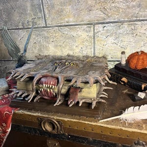 May include: A detailed fantasy book prop with a monstrous mouth and multiple eyes. The book is open, revealing sharp teeth and a red interior. The cover is textured and aged, with a weathered appearance. The prop is displayed on a wooden chest.