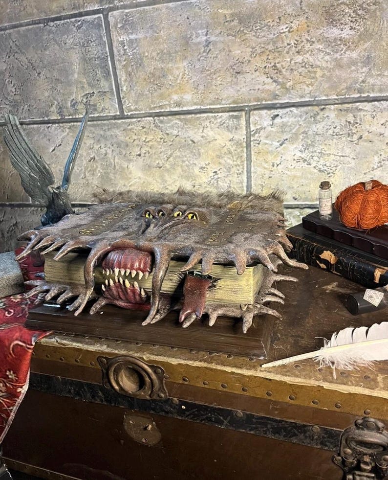 May include: A detailed fantasy book prop with a monstrous mouth and multiple eyes. The book is open, revealing sharp teeth and a red interior. The cover is textured and aged, with a weathered appearance. The prop is displayed on a wooden chest.