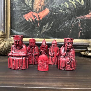 May include: A set of red chess pieces, including kings, queens, bishops, and pawns, are arranged on a dark wooden surface. The pieces have a detailed, carved appearance. The background features a framed painting.