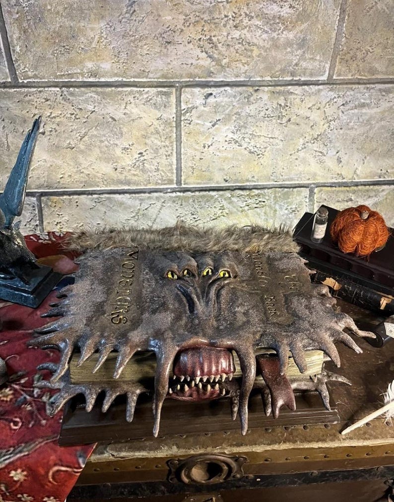 May include: A detailed, open book with a monster-like design. The cover is dark grey with the word "Monsters" and has a furry texture. The book features a large mouth with sharp teeth and multiple yellow eyes.