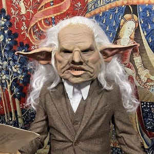 May include: A person wearing a goblin mask with white hair, a brown tweed suit, and a white collared shirt. The mask has large ears and a stern expression. The background features a tapestry with floral and medieval figures.