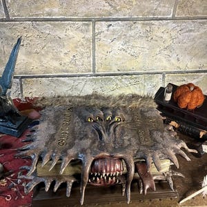May include: A detailed, open book with a monster-like cover. The cover is dark grey with sharp, jagged edges and the word "Monstrorum" is visible. The open mouth reveals sharp teeth. The book is on a wooden surface.