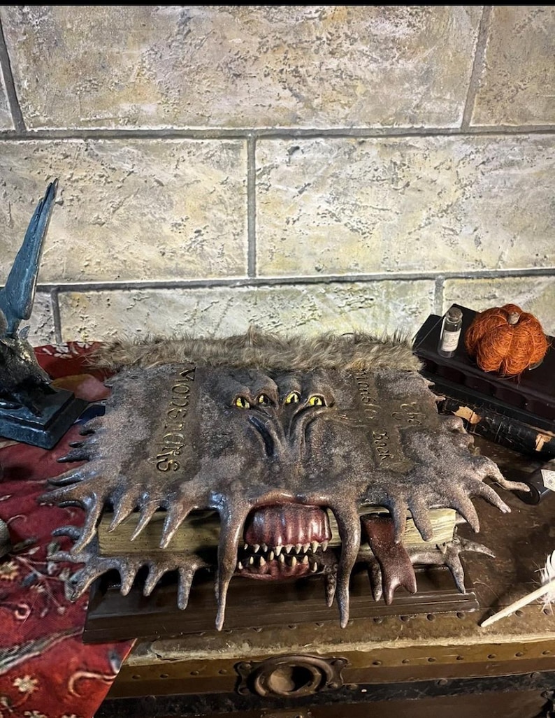May include: A detailed, open book with a monster-like cover. The cover is dark grey with sharp, jagged edges and the word "Monstrorum" is visible. The open mouth reveals sharp teeth. The book is on a wooden surface.