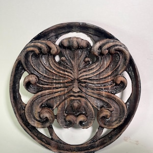May include: A round, brown, wooden wall decor with a carved design of a face with leafy tendrils. The face has a green patina.