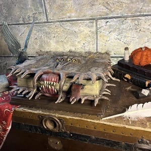 May include: A detailed, open book sculpture with a monster-like mouth and multiple yellow eyes. The book has a fur-like texture and is resting on a wooden chest. A small bottle and a decorative pumpkin are visible in the background.
