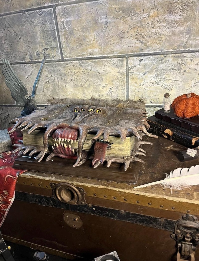 May include: A detailed, open book sculpture with a monster-like mouth and multiple yellow eyes. The book has a fur-like texture and is resting on a wooden chest. A small bottle and a decorative pumpkin are visible in the background.