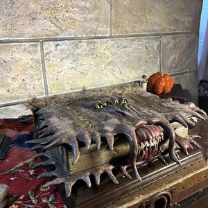 May include: A detailed, handcrafted book with a monster-like design. The cover is dark brown with textured details, featuring a mouth with sharp teeth and multiple yellow eyes. The book is open, revealing aged pages.