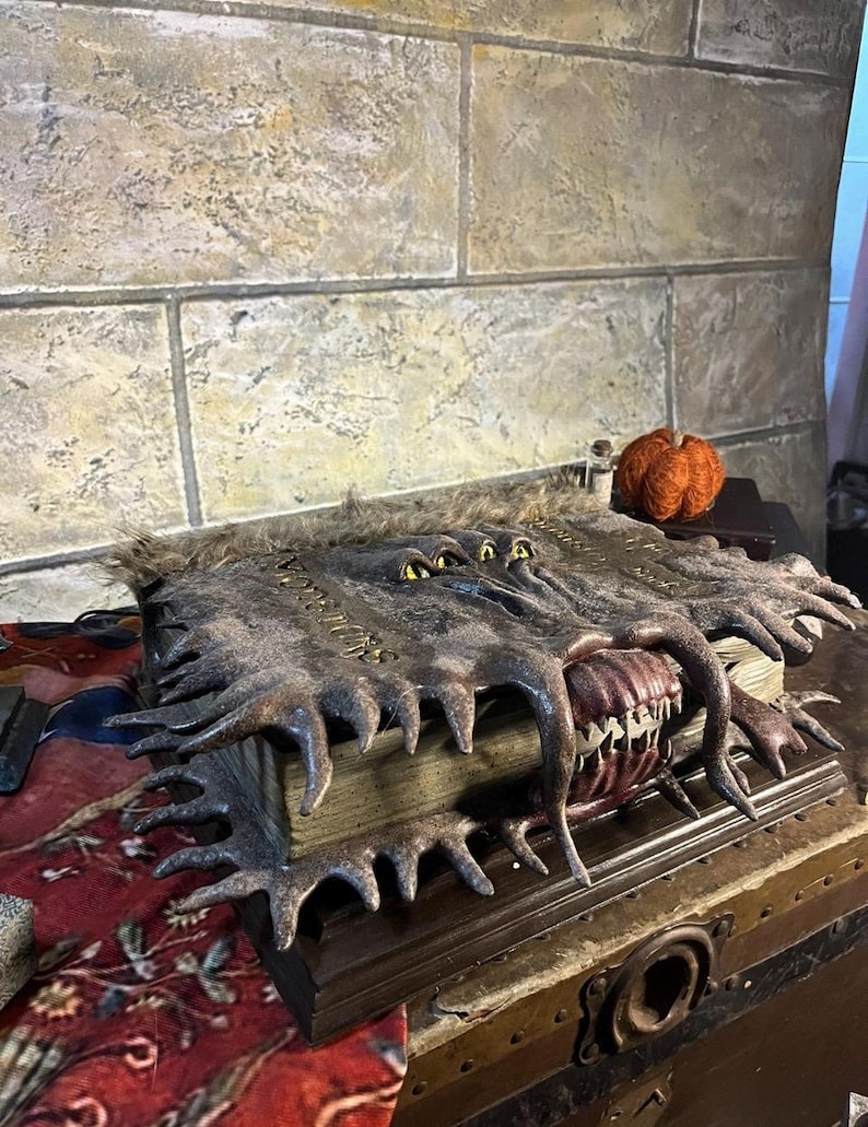 May include: A detailed, handcrafted book with a monster-like design. The cover is dark brown with textured details, featuring a mouth with sharp teeth and multiple yellow eyes. The book is open, revealing aged pages.