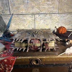 May include: A detailed, handcrafted book sculpture with a monster-like design. The book has a furry cover, multiple eyes, and a large, open mouth with sharp teeth. The sculpture is set on a wooden chest, with other decorative items.
