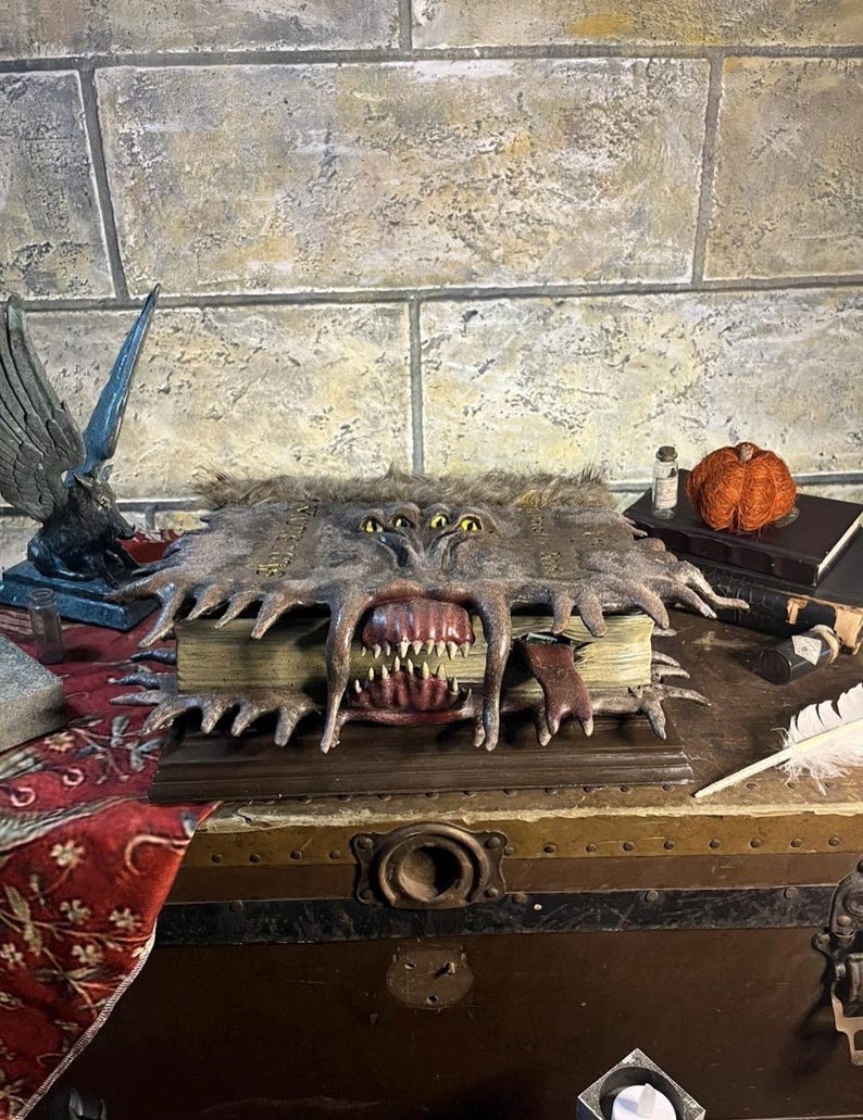 May include: A detailed, handcrafted book sculpture with a monster-like design. The book has a furry cover, multiple eyes, and a large, open mouth with sharp teeth. The sculpture is set on a wooden chest, with other decorative items.