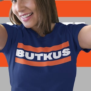 Puede incluir: Una mujer lleva una camiseta azul marino con la palabra "BUTKUS" impresa en letras blancas sobre una franja naranja horizontal. Lleva una gorra de béisbol negra y sonríe.