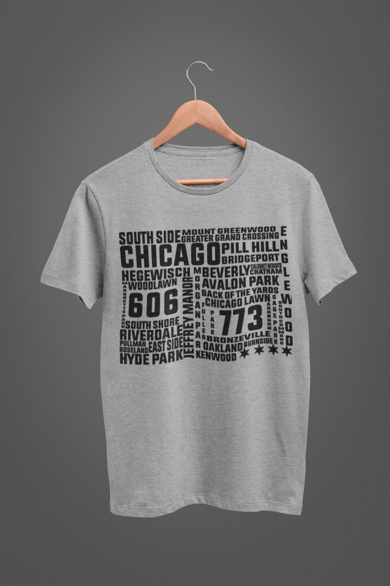 K&ouml;nnte beinhalten: Ein hellgraues T-Shirt mit einem schwarzen Aufdruck, der das Wort "CHICAGO" und verschiedene Stadtteilnamen sowie Postleitzahlen zeigt. Das T-Shirt h&auml;ngt an einem Holzkleiderb&uuml;gel.