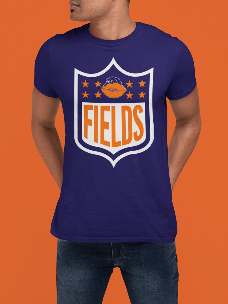May include: A navy blue t-shirt with a white shield design featuring a football and the word "FIELDS" in white letters.