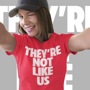May include: A woman wearing a red t-shirt with the text "They're Not Like Us" in white letters. She is smiling and wearing a black baseball cap.