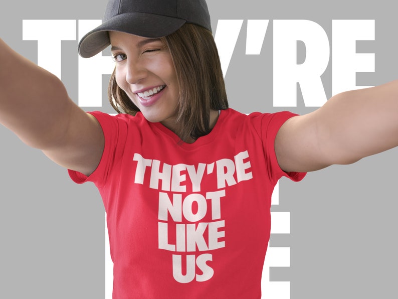 May include: A woman wearing a red t-shirt with the text "They're Not Like Us" in white letters. She is smiling and wearing a black baseball cap.