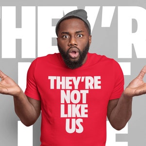 May include: A man wearing a red t-shirt with the text "They're Not Like Us" in white letters. He is looking at the camera with a surprised expression and has his hands raised in the air.