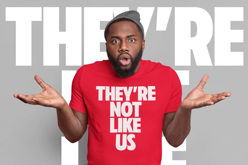 May include: A man wearing a red t-shirt with the text "They're Not Like Us" in white letters. He is looking at the camera with a surprised expression and has his hands raised in the air.