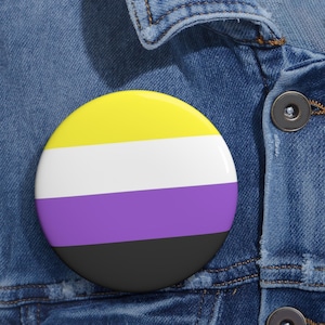 May include: A round button pin featuring the non-binary pride flag. The pin has a yellow top stripe, a white middle stripe, a purple stripe, and a black bottom stripe. The pin is set against a denim jacket.