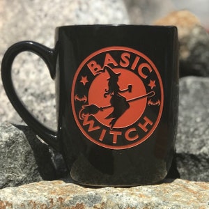 Może przedstawiać: Czarny ceramiczny kubek z dużym uchwytem. Kubek ma okrągły pomarańczowy wzór z sylwetką wiedźmy na miotle i napisem "BASIC WITCH" w pomarańczowych literach. Kubek stoi na skale.