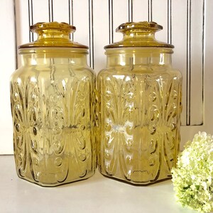 May include: Two amber-colored glass jars with lids. The jars have a square shape and are decorated with a repeating floral pattern. The lids are also amber and have a decorative knob. The jars are on a white surface.