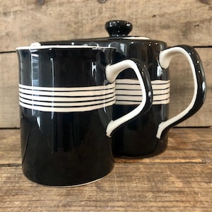 May include: A black ceramic mug and a matching teapot with a lid, both featuring a series of thin white horizontal stripes around the upper section. Each piece has a curved white handle and a glossy black finish on the exterior surface.