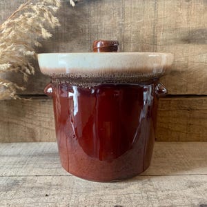 May include: A vintage, glazed ceramic crock with a lid. The crock is a deep reddish-brown colour with a lighter tan rim and lid. It has two small handles on the sides. The background is a rustic wooden surface.