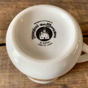 K&ouml;nnte beinhalten: Wei&szlig;e Disney-Tasse aus Keramik mit Henkel. Der Boden der Tasse zeigt die Logos von Disneyland und Walt Disney World mit dem Text "Designed and made for Disneyland Walt Disney World" und "Made in Japan".