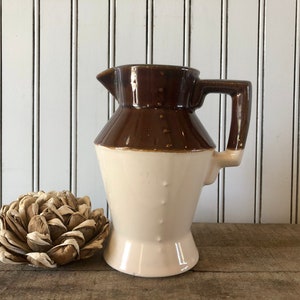 May include: A vintage brown and cream-colored ceramic pitcher with a handle and spout. The pitcher has a two-tone design, with a dark brown top and a cream-colored base. A decorative dried flower is to the left of the pitcher.