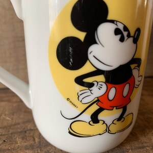K&ouml;nnte beinhalten: Wei&szlig;e Keramiktasse mit einer Mickey Mouse-Illustration. Die Zeichentrickfigur ist in ihrem klassischen Outfit dargestellt, mit schwarzen Ohren, einem wei&szlig;en Gesicht, roten Shorts und gelben Schuhen. Die Tasse hat einen gebogenen Griff und einen gelben Hintergrund.