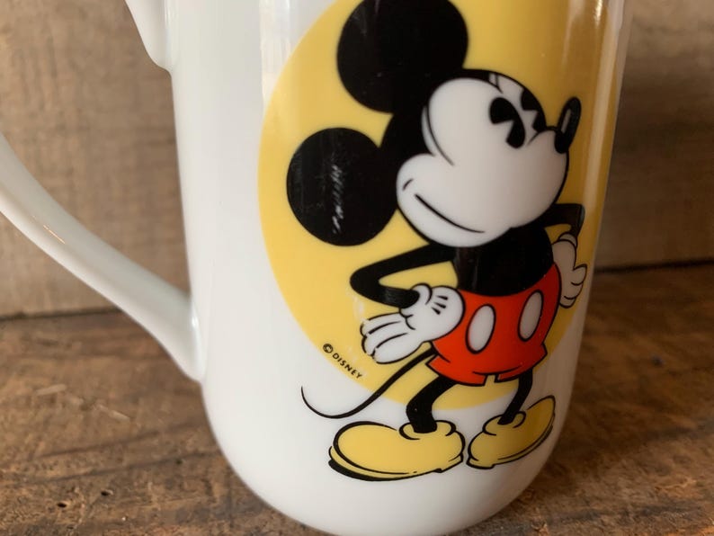 K&ouml;nnte beinhalten: Wei&szlig;e Keramiktasse mit einer Mickey Mouse-Illustration. Die Zeichentrickfigur ist in ihrem klassischen Outfit dargestellt, mit schwarzen Ohren, einem wei&szlig;en Gesicht, roten Shorts und gelben Schuhen. Die Tasse hat einen gebogenen Griff und einen gelben Hintergrund.